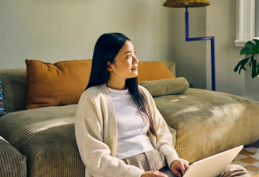 Woman sitting on a couch, looking to the side while using a laptop on her lap.
